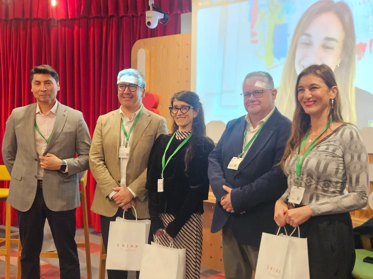 Cinco participantes de la conferencia posan con bolsas de Eniax; al fondo, en pantalla, una expositora conectada en línea.