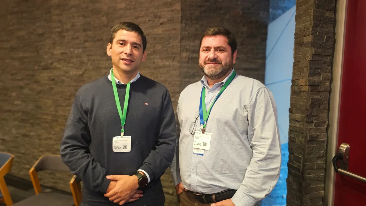 Dos asistentes con credencial posan ante un muro de piedra oscura.